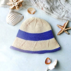 May include: A beige knitted bucket hat with a blue stripe around the crown. The hat is on a white surface with seashells and a starfish.