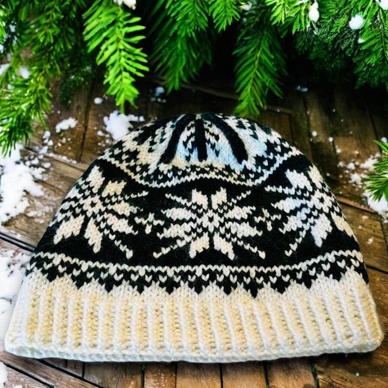 May include: A knitted winter hat with a snowflake pattern. The hat features a cream-colored brim and a black and cream snowflake design on the crown. The hat is displayed on a wooden surface with evergreen branches in the background.