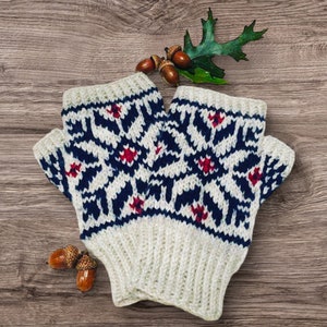 May include: A pair of white knitted fingerless gloves with a blue and red geometric pattern. The gloves are on a wooden surface with acorns.