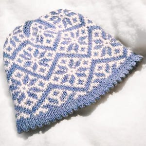 以下が含まれることがあります： 雪の結晶模様の青と白のニット帽。帽子の縁は折り返されており、柔らかな糸でできています。