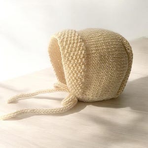 May include: A knitted baby bonnet in a light beige color with a tied bow at the back.