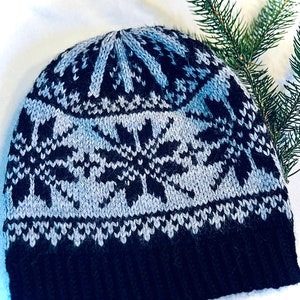 May include: A knitted winter hat with a snowflake design. The hat features a black brim, a light gray and black snowflake pattern, and a light gray top. The hat is made of a soft, warm material, perfect for cold weather.