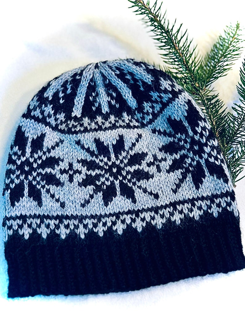 May include: A knitted winter hat with a snowflake design. The hat features a black brim, a light gray and black snowflake pattern, and a light gray top. The hat is made of a soft, warm material, perfect for cold weather.