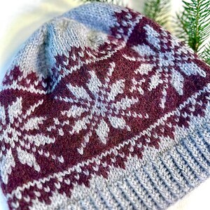 May include: A gray and burgundy knit hat with a snowflake pattern. The hat features a ribbed brim and a detailed design across the crown. The color scheme includes shades of gray and burgundy, creating a warm, wintery aesthetic.