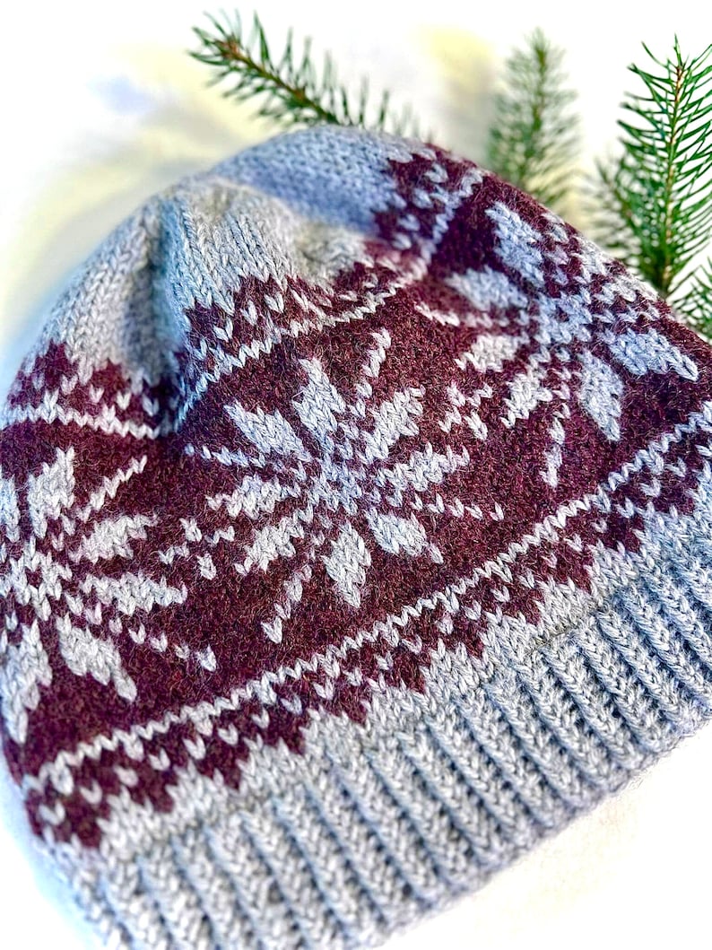 May include: A gray and burgundy knit hat with a snowflake pattern. The hat features a ribbed brim and a detailed design across the crown. The color scheme includes shades of gray and burgundy, creating a warm, wintery aesthetic.
