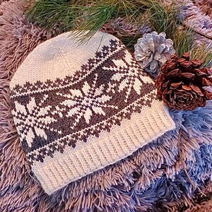 May include: A cream-colored knit beanie with a snowflake pattern in dark gray and white. The hat features a ribbed brim and a detailed geometric design. The hat is displayed on a fluffy, textured surface with pine needles and pine cones.