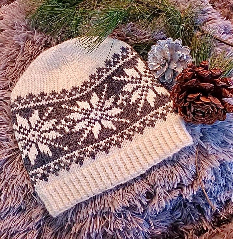 May include: A cream-colored knit beanie with a snowflake pattern in dark gray and white. The hat features a ribbed brim and a detailed geometric design. The hat is displayed on a fluffy, textured surface with pine needles and pine cones.