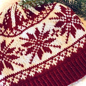 May include: A knitted winter hat in burgundy and cream. The hat features a repeating snowflake pattern in cream against a burgundy background. The brim is a solid burgundy color. A sprig of evergreen is in the background.