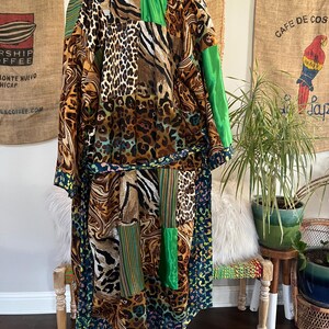 May include: A long, patchwork kimono-style robe featuring a mix of animal prints, including leopard and zebra patterns, with green and blue accents. The robe has a belt and is displayed on a stand. The background includes burlap coffee sacks and plants.