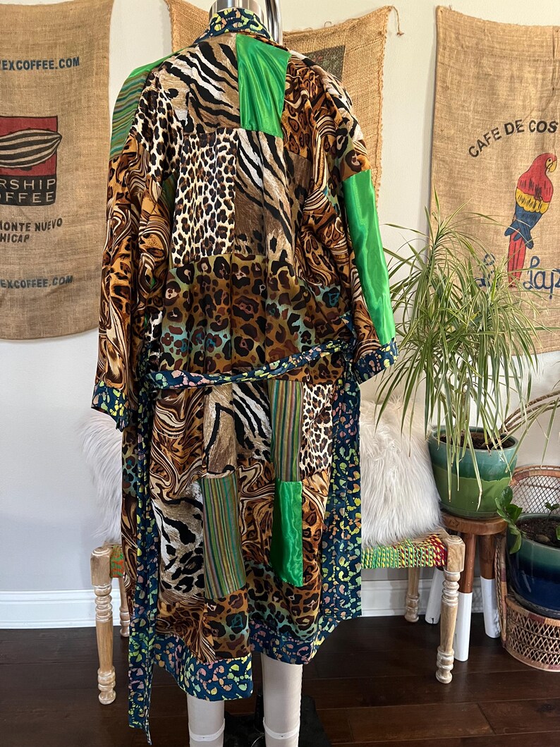 May include: A long, patchwork kimono-style robe featuring a mix of animal prints, including leopard and zebra patterns, with green and blue accents. The robe has a belt and is displayed on a stand. The background includes burlap coffee sacks and plants.