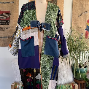 May include: A patchwork kimono-style jacket featuring a variety of fabrics including green patterned cotton, solid purple panels, denim pockets with braided trim, and a denim closure tab with a button. The garment has wide sleeves and an open front.