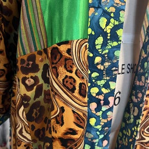 May include: A patchwork garment featuring a variety of patterns and colors. The design includes sections of green, leopard print, striped fabric, and a blue batik-style pattern. The garment is draped over a mannequin.