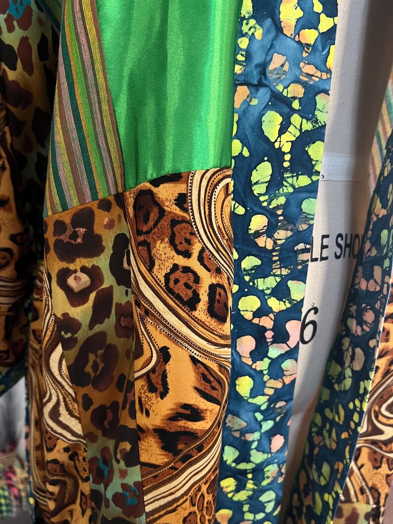 May include: A patchwork garment featuring a variety of patterns and colors. The design includes sections of green, leopard print, striped fabric, and a blue batik-style pattern. The garment is draped over a mannequin.