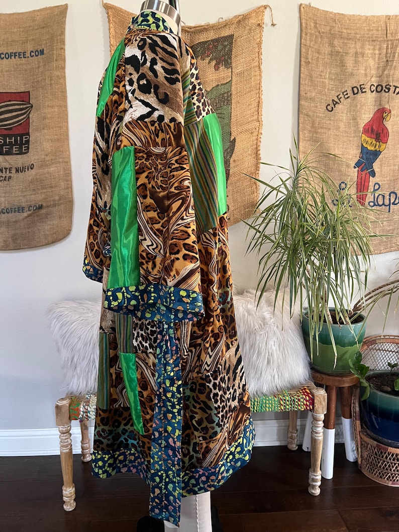 May include: A long, patchwork robe with a vibrant mix of patterns and colors. The robe features leopard print, green satin panels, and floral accents. The robe is displayed on a mannequin, showcasing its flowing design and unique style.