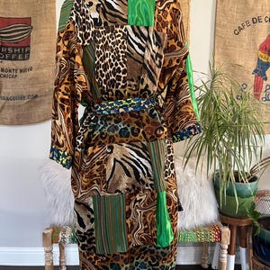 May include: A long, patchwork robe featuring a mix of animal prints, including leopard and zebra patterns, with green and striped accents. The robe has a matching belt and is displayed on a mannequin. The robe's hemline and cuffs have a dark blue and yellow floral design.