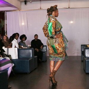 May include: A model in a patterned dress with green accents and a matching belt walks on a runway. The dress features a mix of animal print and floral designs. The model is wearing matching patterned high heels. The audience is seated in blue booths.