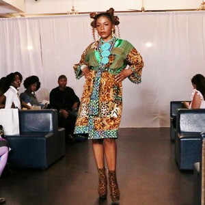 May include: A woman modeling a colorful patterned dress with a green top and a brown and tan skirt. The dress has a belt and is paired with matching patterned ankle boots. The model is standing on a runway in front of an audience.