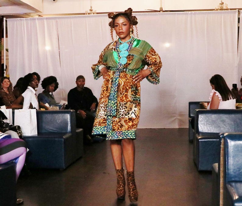 May include: A woman modeling a colorful patterned dress with a green top and a brown and tan skirt. The dress has a belt and is paired with matching patterned ankle boots. The model is standing on a runway in front of an audience.