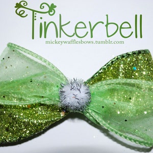 Peut inclure: Un nœud vert en tissu brillant et transparent avec un pompon blanc au centre. Le nœud est décoré de paillettes dorées. Le texte "Tinkerbell" est écrit en cursive verte au-dessus du nœud. Le texte "mickeywafflesbows.tumblr.com" est écrit sous le nœud.