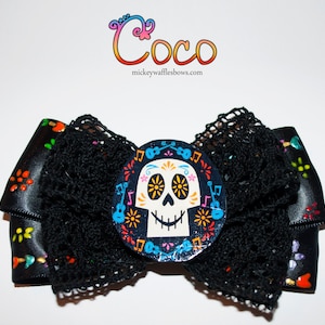May include: Black and white bow with a colourful sugar skull design in the centre. The skull has a floral pattern and is surrounded by musical notes and guitars. The bow is made of black satin ribbon and black lace.