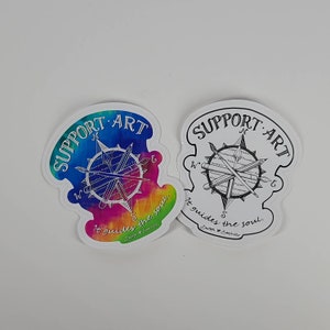 May include: Two die-cut stickers with a compass design. One features a colorful tie-dye background with the words "SUPPORT ART" and "It guides the soul." The other is black and white with the same text.