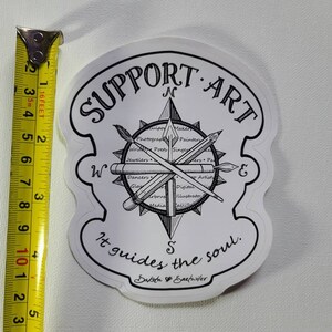 May include: A white sticker with black text and illustrations. The sticker features a compass design with the words "SUPPORT ART" at the top. The compass includes illustrations of art tools and the text "It guides the soul."