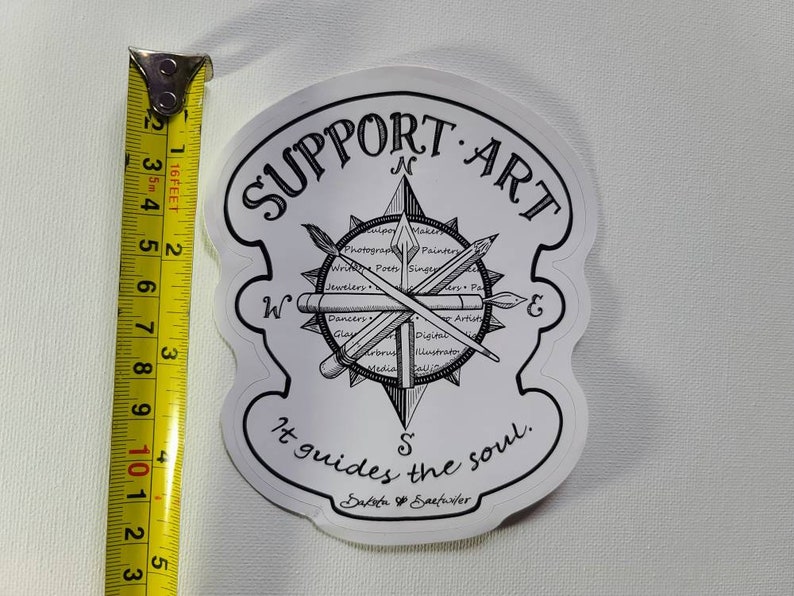 May include: A white sticker with black text and illustrations. The sticker features a compass design with the words "SUPPORT ART" at the top. The compass includes illustrations of art tools and the text "It guides the soul."