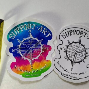 May include: Two stickers with a compass design and the text "SUPPORT ART" and "It guides the soul." One sticker has a colorful watercolor background, the other is black and white. The stickers are approximately 3 inches (7.6 cm) wide.