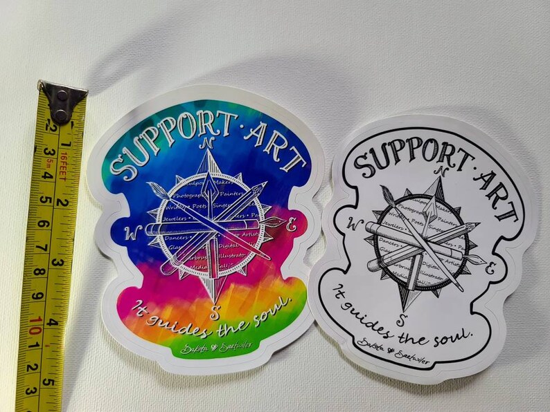 May include: Two stickers with a compass design and the text "SUPPORT ART" and "It guides the soul." One sticker has a colorful watercolor background, the other is black and white. The stickers are approximately 3 inches (7.6 cm) wide.