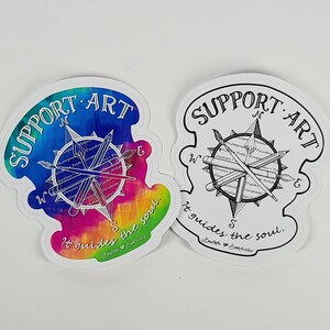May include: Two die-cut stickers with a compass design. One features a watercolor rainbow background, the other is black and white. Both say "SUPPORT ART" and "It guides the soul."