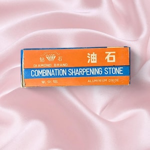 May include: A rectangular box for a combination sharpening stone. The box is orange and blue with text that reads "DIAMOND BRAND COMBINATION SHARPENING STONE ALUMINIUM OXIDE". The box is on a pink satin surface.