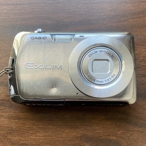 May include: A silver Casio Exilim digital camera with a metallic finish. The front features the brand name Casio, the model name Exilim, and a lens with the text Exilim Optical 3x, f=6.3-18.9mm, 1:3.1-5.6. A small flash unit is located at the top.