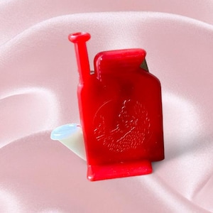 May include: A red watering can-shaped pin with a white spout. The pin features a detailed embossed design on the front. The pin is set against a pink background.