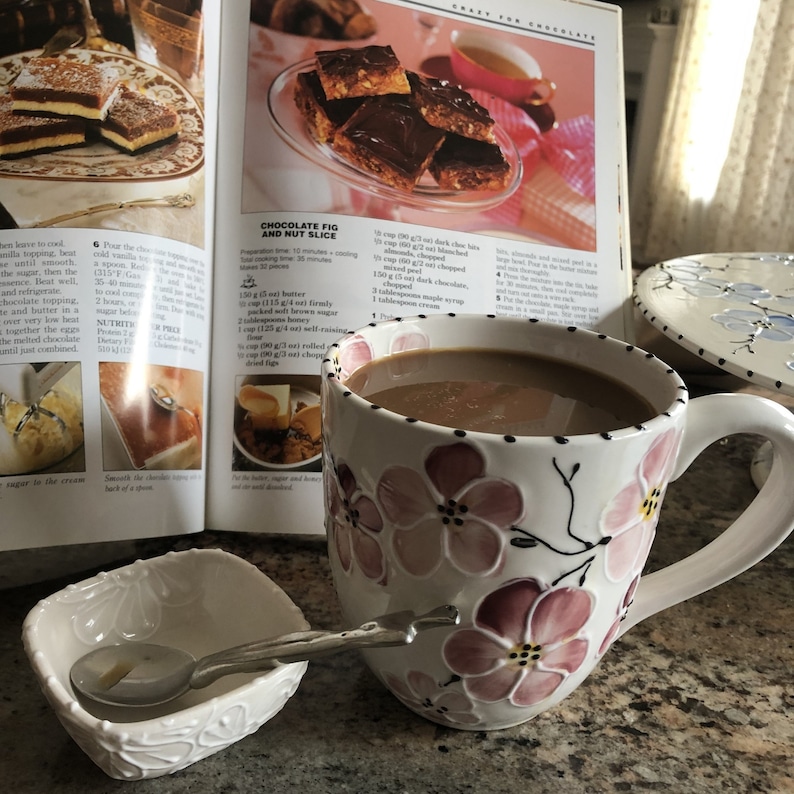 May include: A white ceramic mug with pink floral designs is filled with coffee. The mug is sitting on a table next to a cookbook open to a recipe for chocolate fig and nut slice. The recipe includes ingredients such as butter, brown sugar, rolled oats, and chopped figs.
