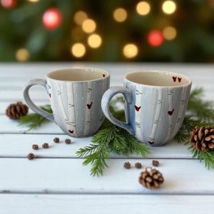 May include: Two ceramic mugs with a light blue birch tree design and red heart accents. The mugs have curved handles. The mugs are surrounded by pine needles and pine cones, creating a festive holiday theme.
