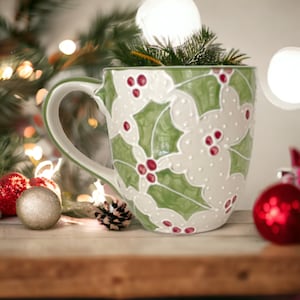 May include: A green and white ceramic mug with a holly leaf design and red berries. The mug is sitting on a wooden surface with Christmas decorations and lights in the background.