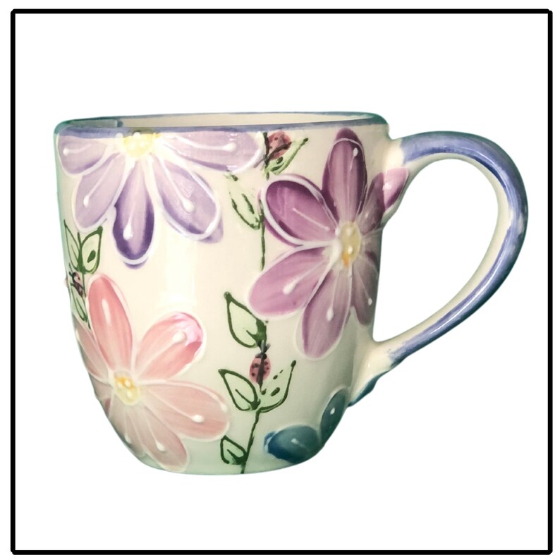 May include: A white ceramic mug with a floral design. The mug features pink, purple and blue flowers with green leaves. The handle is blue.