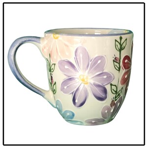 May include: A white ceramic mug with a blue handle and a floral design. The flowers are painted in shades of pink, purple, blue, and green.