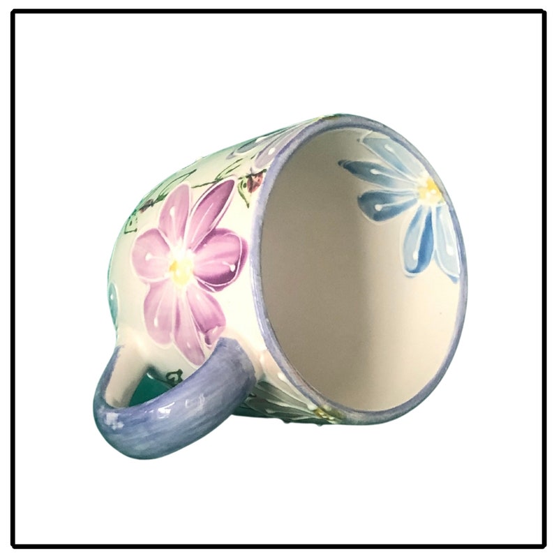 May include: A white ceramic mug with a blue handle and floral design. The flowers are painted in shades of purple, blue, and green.