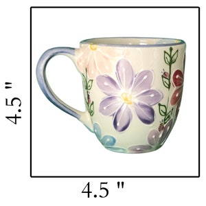 May include: A white ceramic mug with a floral design. The mug has a blue handle and is decorated with pink, purple, and blue flowers. The mug is 4.5 inches tall and 4.5 inches wide.