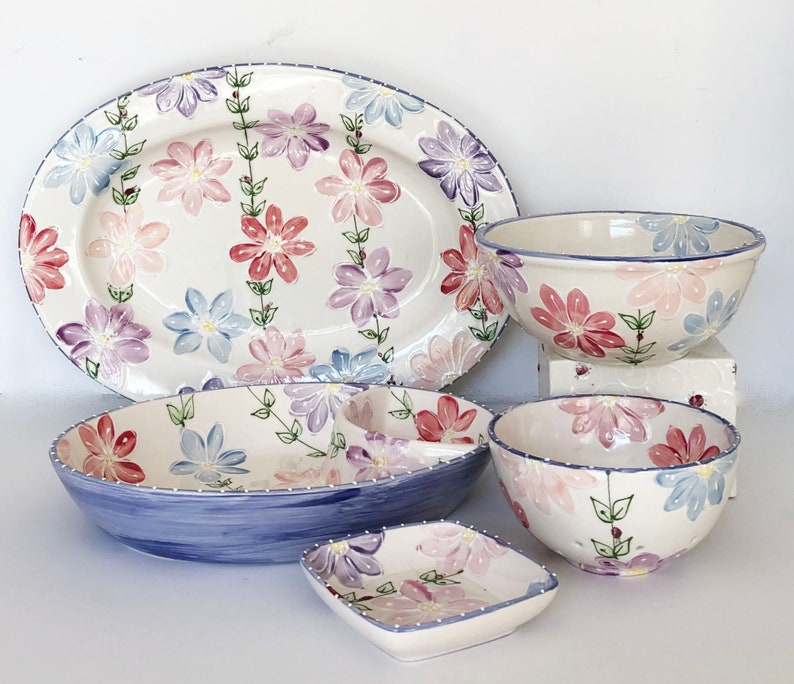 May include: A set of five ceramic dishes featuring a floral design in shades of pink, purple, and blue. The set includes a large oval platter, two bowls, and two smaller dishes.
