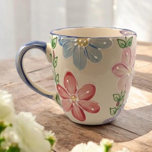 May include: A white ceramic mug with a blue handle and a floral design. The flowers are pink, blue, and green. The mug is sitting on a wooden table.
