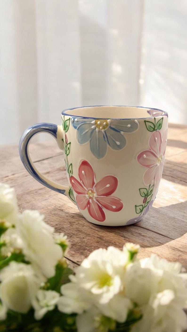 May include: A white ceramic mug with a blue handle and a floral design. The flowers are pink, blue, and green. The mug is sitting on a wooden table.