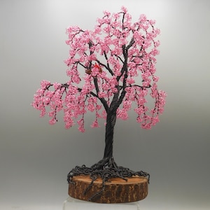 May include: A handcrafted wire tree sculpture with a dark trunk and branches, adorned with pink beaded blossoms. The tree sits on a wooden base. A small red bird is nestled in the branches. The sculpture is approximately 10 inches tall.