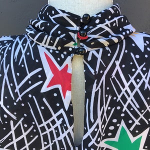 Puede incluir: Blusa de estampado blanco y negro con cuello alto y abertura de ojo de cerradura en la parte delantera. La blusa tiene estrellas rojas, verdes y blancas. La blusa tiene tres botones negros en el cuello.