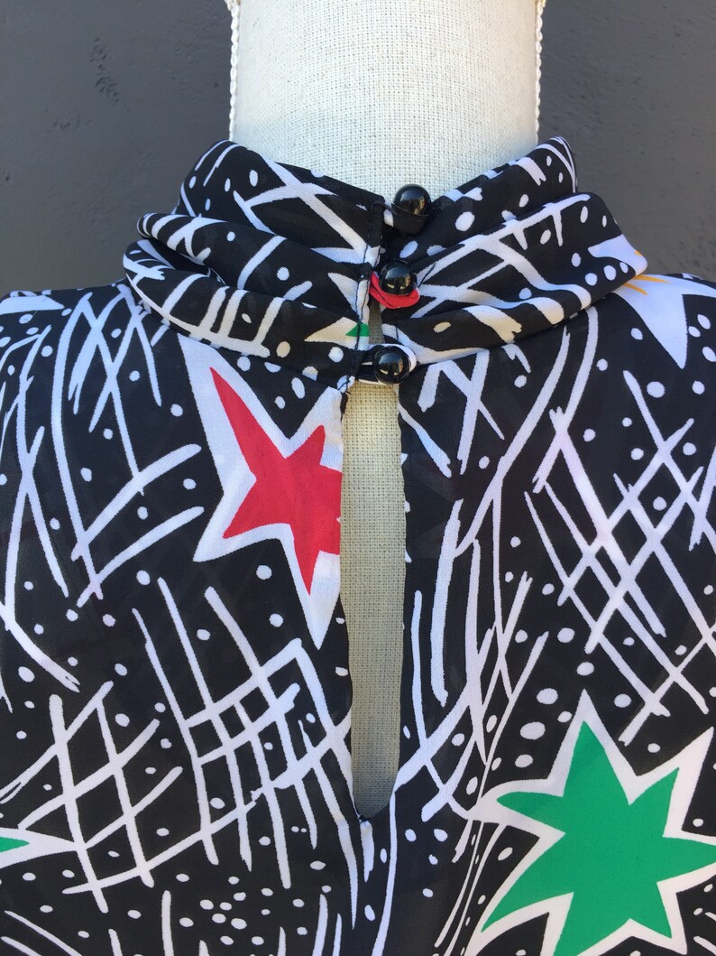 Puede incluir: Blusa de estampado blanco y negro con cuello alto y abertura de ojo de cerradura en la parte delantera. La blusa tiene estrellas rojas, verdes y blancas. La blusa tiene tres botones negros en el cuello.