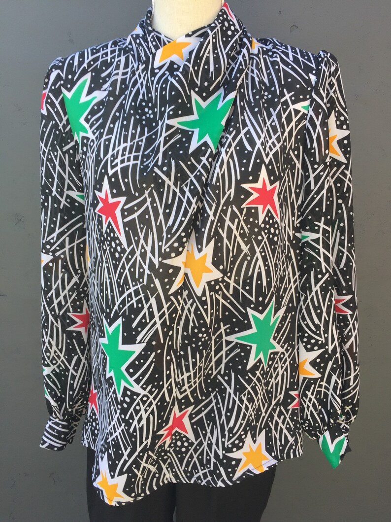 Puede incluir: Una blusa negra de manga larga con un estampado de estrellas de colores. Las estrellas son rojas, verdes, amarillas y blancas. El fondo es negro con l&iacute;neas blancas.