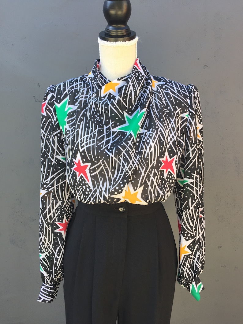 Puede incluir: Una blusa de estampado blanco y negro con estrellas de colores. La blusa tiene cuello alto y mangas largas. La blusa se combina con pantalones negros.