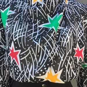 Puede incluir: Una blusa de estampado blanco y negro con estrellas rojas, verdes y amarillas. La blusa tiene cuello alto y mangas largas.