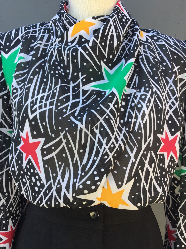 Puede incluir: Una blusa de estampado blanco y negro con estrellas rojas, verdes y amarillas. La blusa tiene cuello alto y mangas largas.